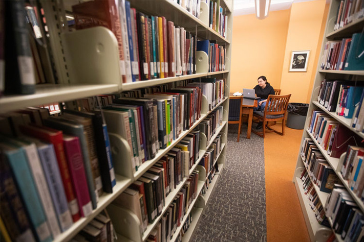 Rows of bookshelves in a library with a person working on a laptop at a table.