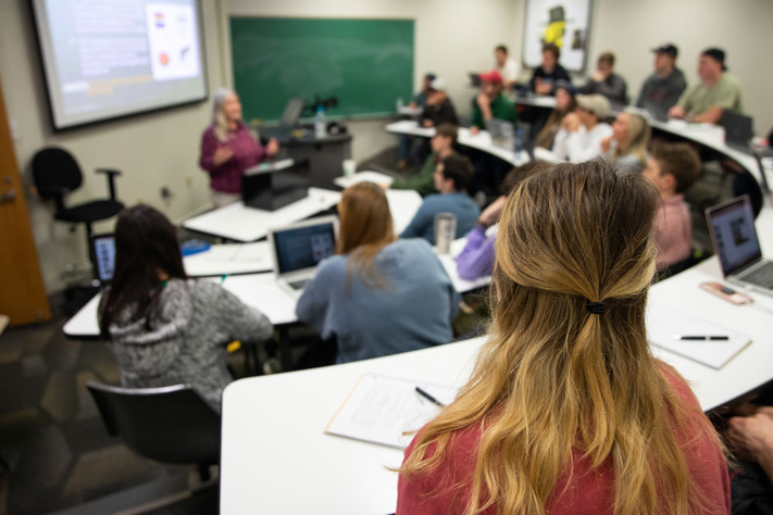 Classroom setting with students seated at desks, listening to a lecture with slides projected.