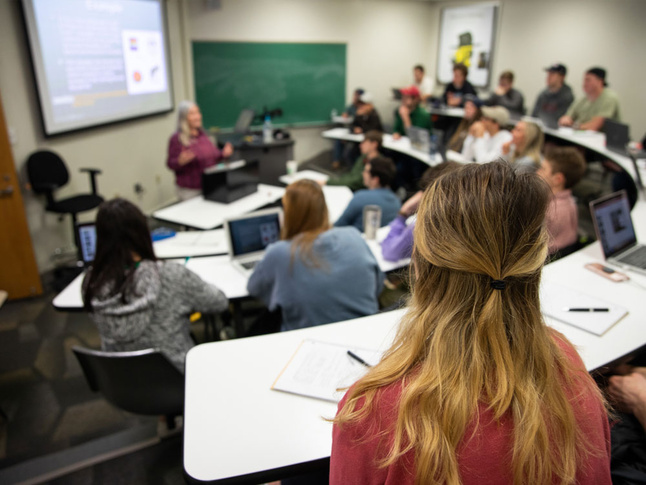 Classroom setting with students seated at desks, listening to a lecture with slides projected.
